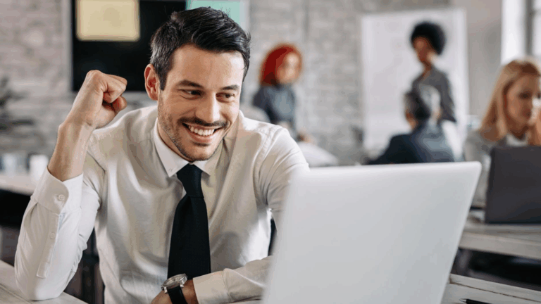 A young professional man in a white shirt and black tie smiles and pumps his fist in success while working on a laptop in a modern office. The image conveys the positive impact of cloud-based communication and VoIP systems in a collaborative, cost-effective telecom environment.