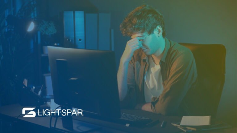 Frustrated man in a dark office holds his forehead while looking at a desktop computer screen. The scene suggests the stress of outdated systems and the need for business network modernization and infrastructure resilience.