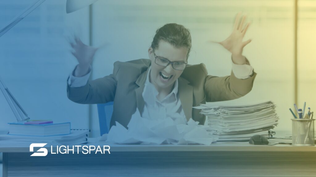 A frustrated woman in business attire throws papers in the air at her cluttered desk, surrounded by stacks of documents and office supplies. The Lightspar logo appears in the corner. The image conveys the chaos that unified communications can help solve, highlighting the need for UCaaS solutions in modern workplaces.