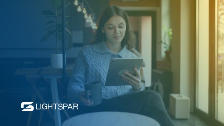 A woman sits in a modern, cozy workspace, smiling while using a tablet and holding a coffee mug. The LightSpar logo appears in the corner, representing the flexibility and mobility benefits of business VoIP.